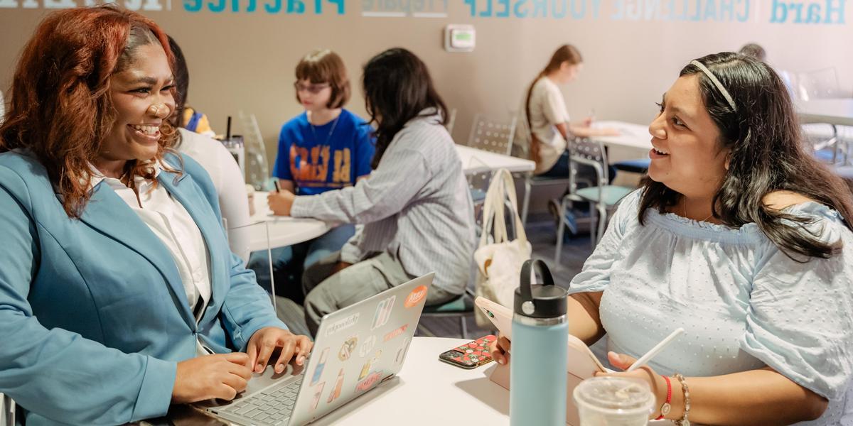 Two students chat and smile while working on a laptop and a tablet